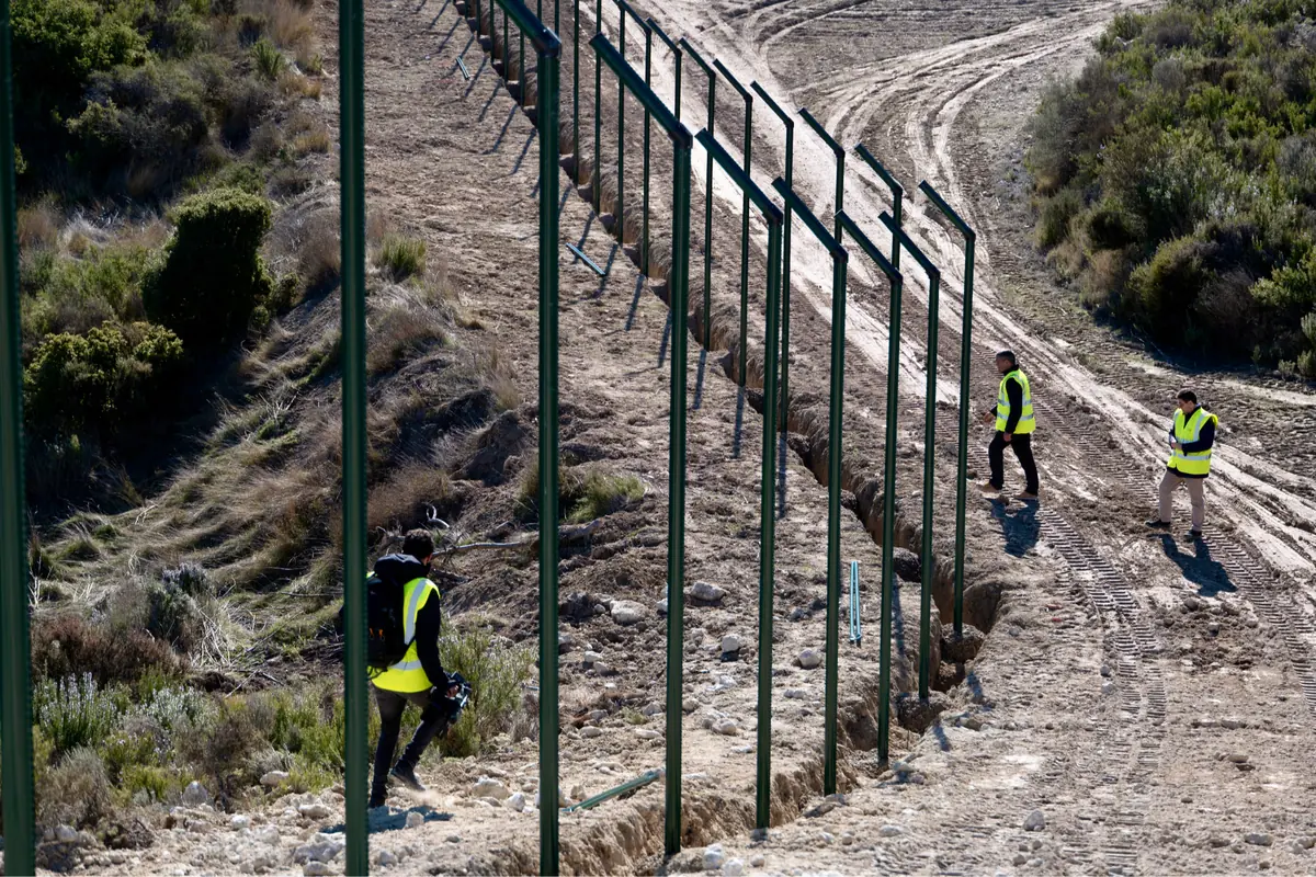 Trabajadores con chalecos reflectantes inspeccionan la instalación de una valla metálica en un terreno rural de tierra y matorrales