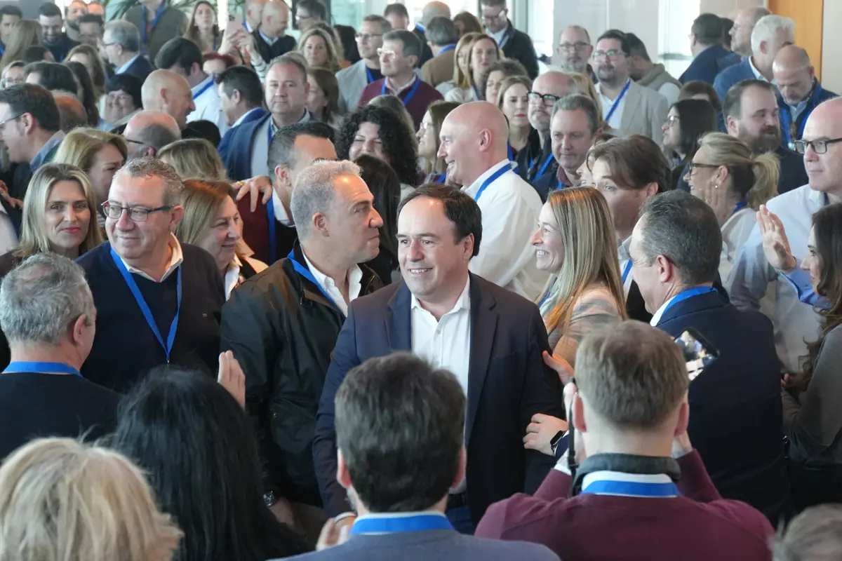Hombre de traje oscuro y camisa blanca sonríe mientras camina entre una multitud de personas con acreditaciones azules en un evento o conferencia