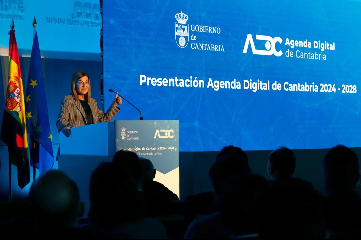 Conferencia de presentación de la Agenda Digital de Cantabria 2024-2028 con una ponente hablando en un escenario junto a las banderas de España y la Unión Europea frente a un público sentado