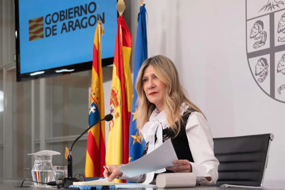 Mujer rubia sentada en una sala de prensa del Gobierno de Aragón con banderas oficiales detrás mientras sostiene unos documentos frente a un micrófono Mujer rubia sentada en una sala de prensa del Gobierno de Aragón con banderas oficiales detrás mientras sostiene unos documentos frente a un micrófono