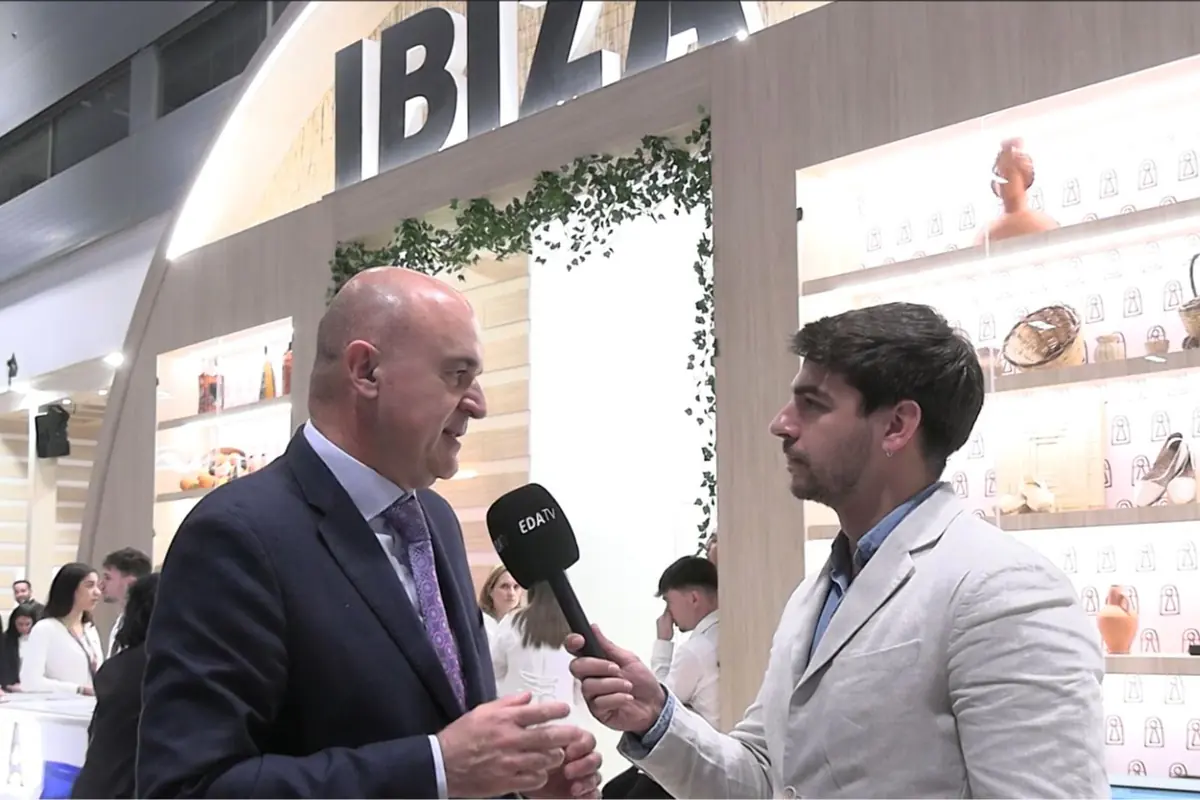 Entrevista en un stand de feria de turismo de Ibiza con un periodista sosteniendo un micrófono de EDATV frente a un hombre trajeado mientras varias personas trabajan y exponen productos en el fondo