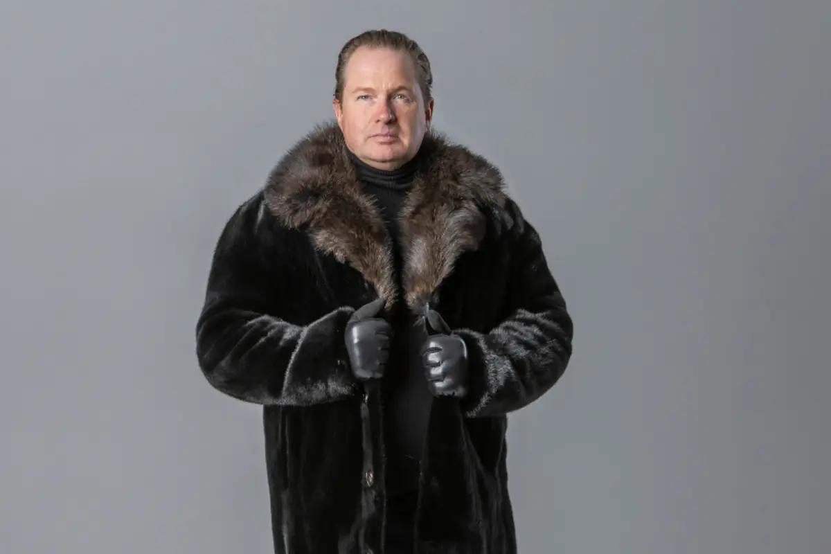 Hombre de pie con abrigo de piel oscuro y guantes negros sobre fondo gris