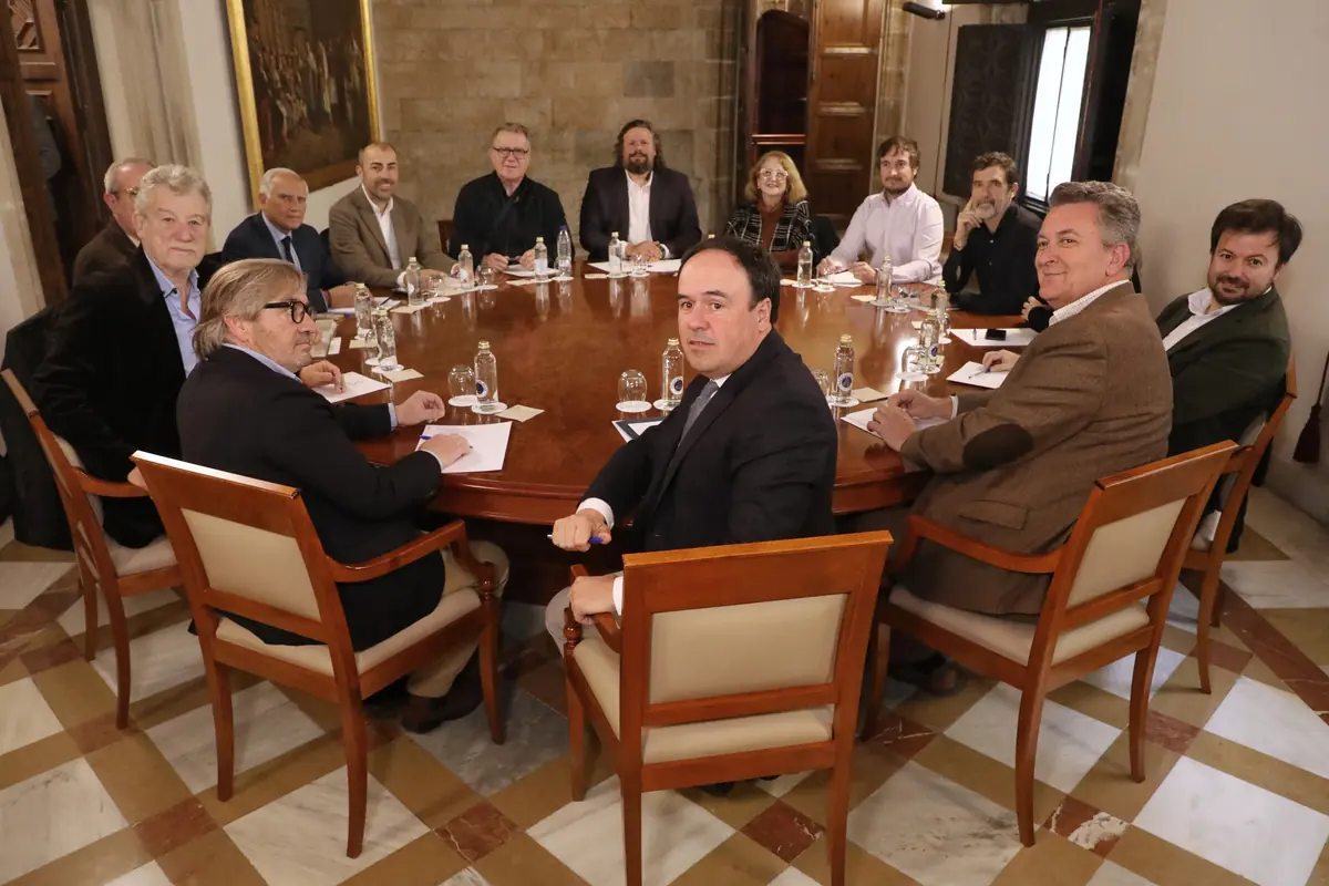Reunión formal de doce personas sentadas alrededor de una gran mesa redonda de madera en una sala elegante con paredes de piedra y cuadros al fondo
