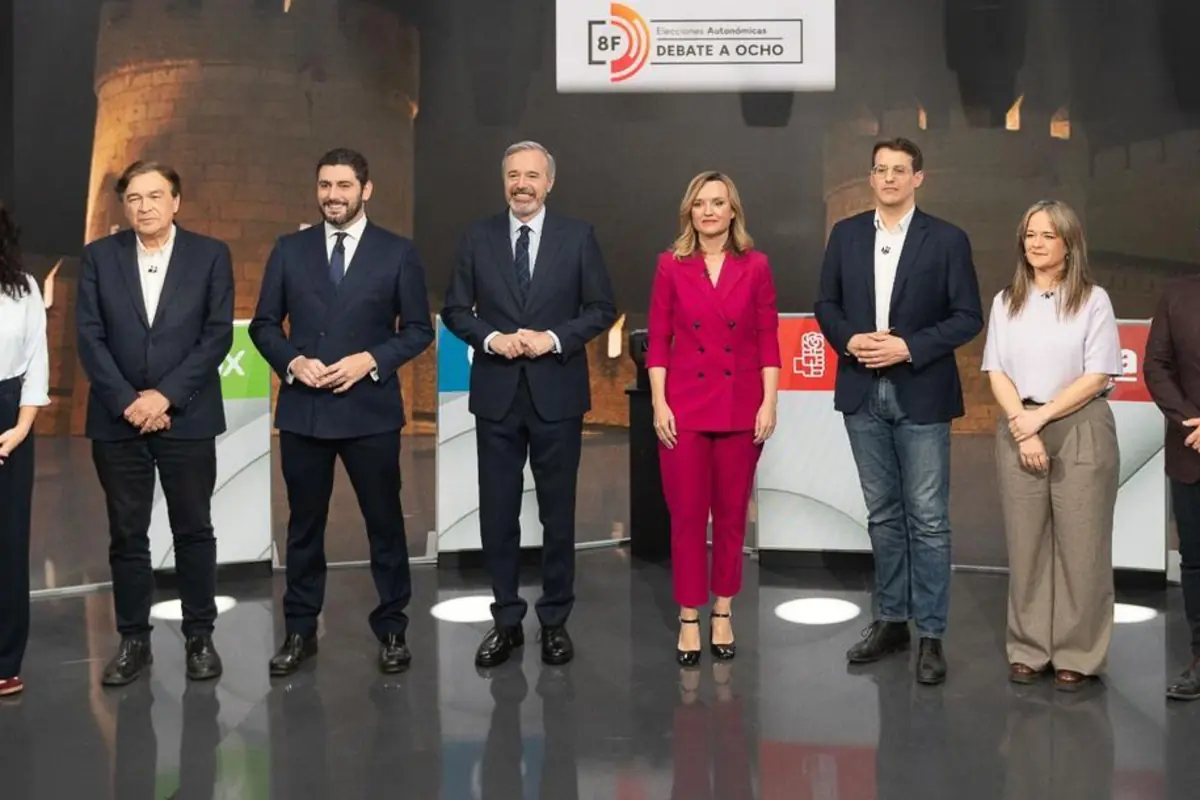 Ocho personas de pie en un plató de televisión posan antes de un debate electoral, alineadas frente a atriles con logotipos de partidos políticos y un cartel que indica debate a ocho