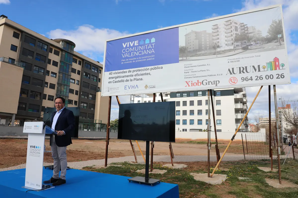Hombre hablando en un atril sobre una tarima azul frente a un solar urbano con un gran cartel publicitario que anuncia la construcción de 40 viviendas de protección pública en Castelló de la Plana