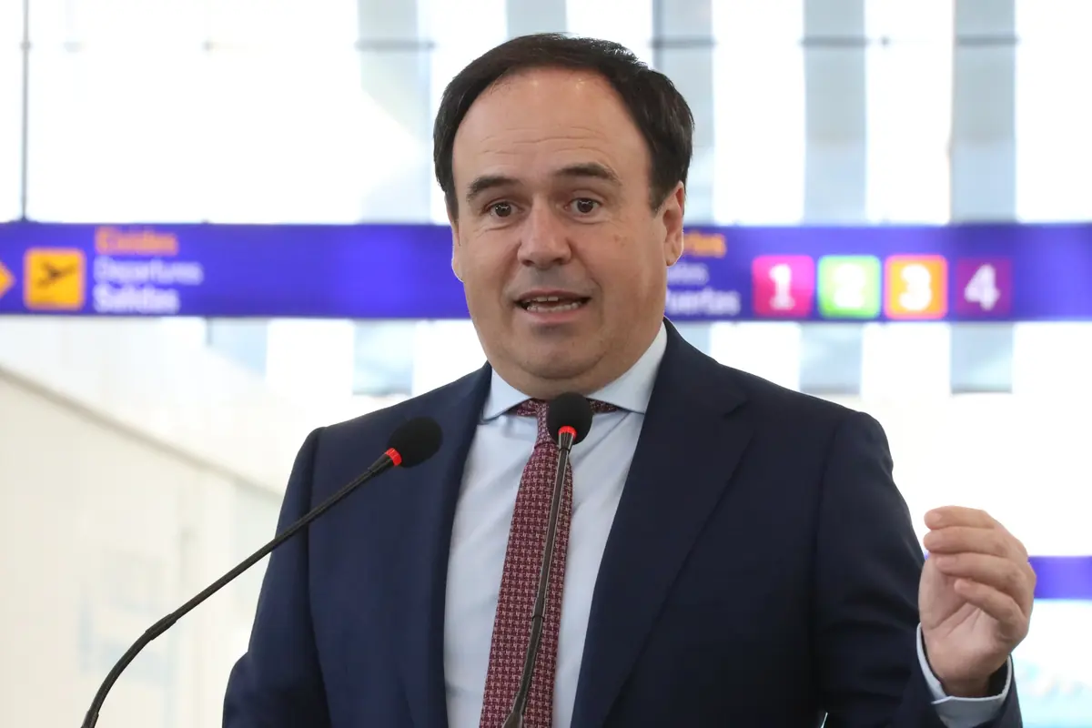 Hombre con traje y corbata hablando frente a dos micrófonos en el interior de un aeropuerto con carteles de salidas al fondo