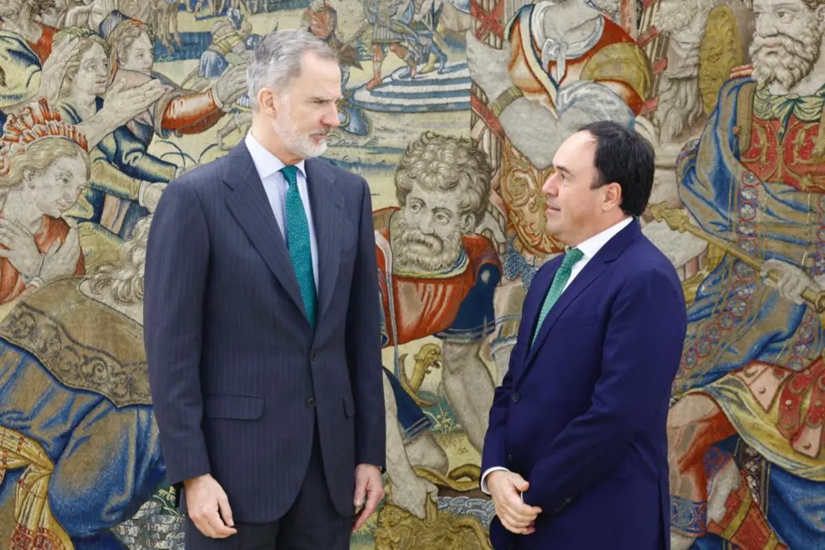 Dos hombres con traje oscuro y corbata verde conversan de pie frente a un gran tapiz histórico de colores vivos