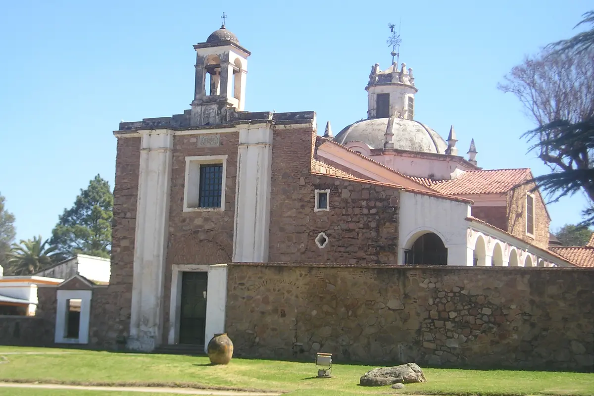 Iglesia colonial de piedra con cúpula y campanario rodeada de un muro bajo y césped en un día soleado