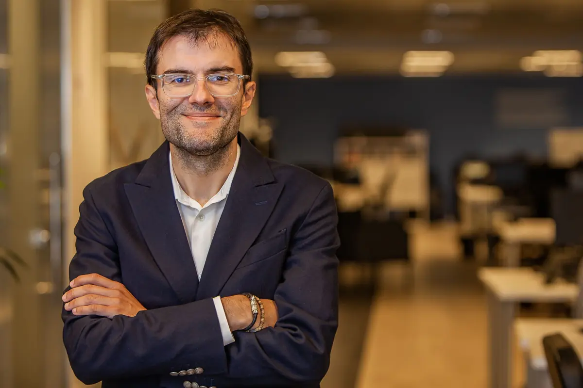 Hombre con gafas y traje azul sonriendo con los brazos cruzados en una oficina moderna desenfocada al fondo