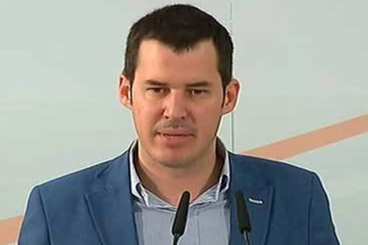 Hombre de mediana edad con chaqueta azul hablando frente a dos micrófonos sobre un fondo claro