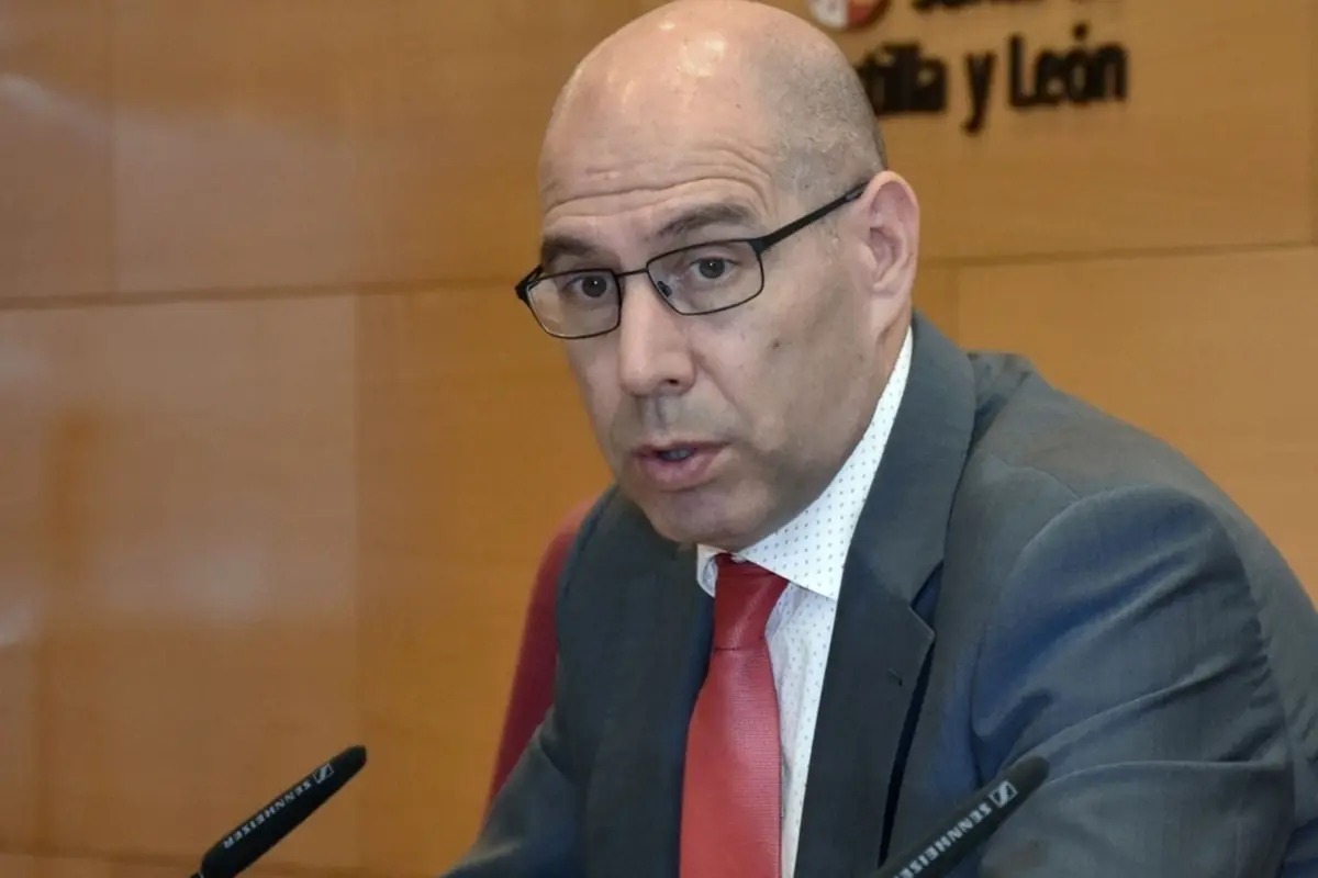 Hombre calvo con gafas y traje gris con corbata roja hablando frente a micrófonos en una sala de conferencias