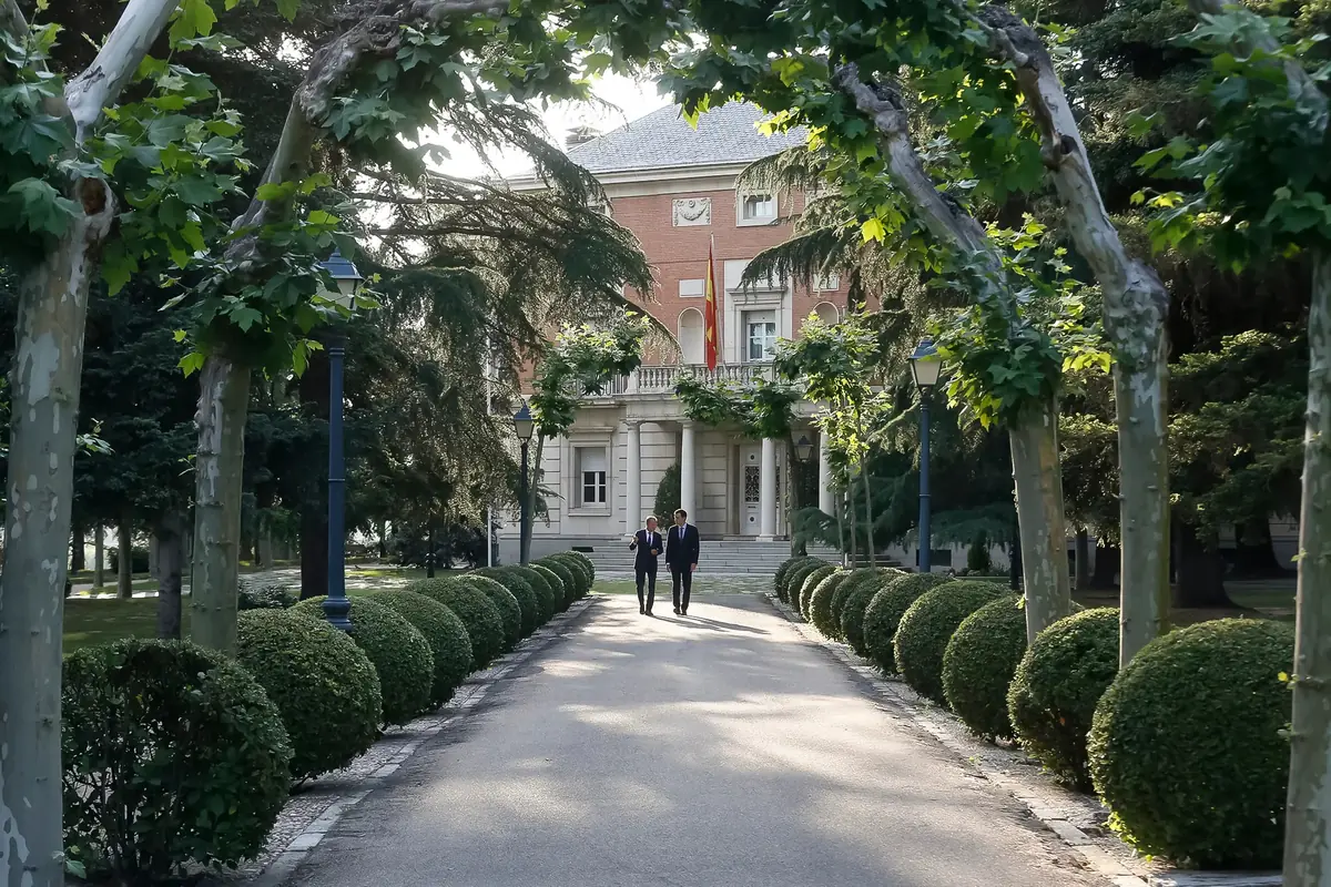 Dos personas caminando por una avenida arbolada que conduce a un edificio oficial con bandera española
