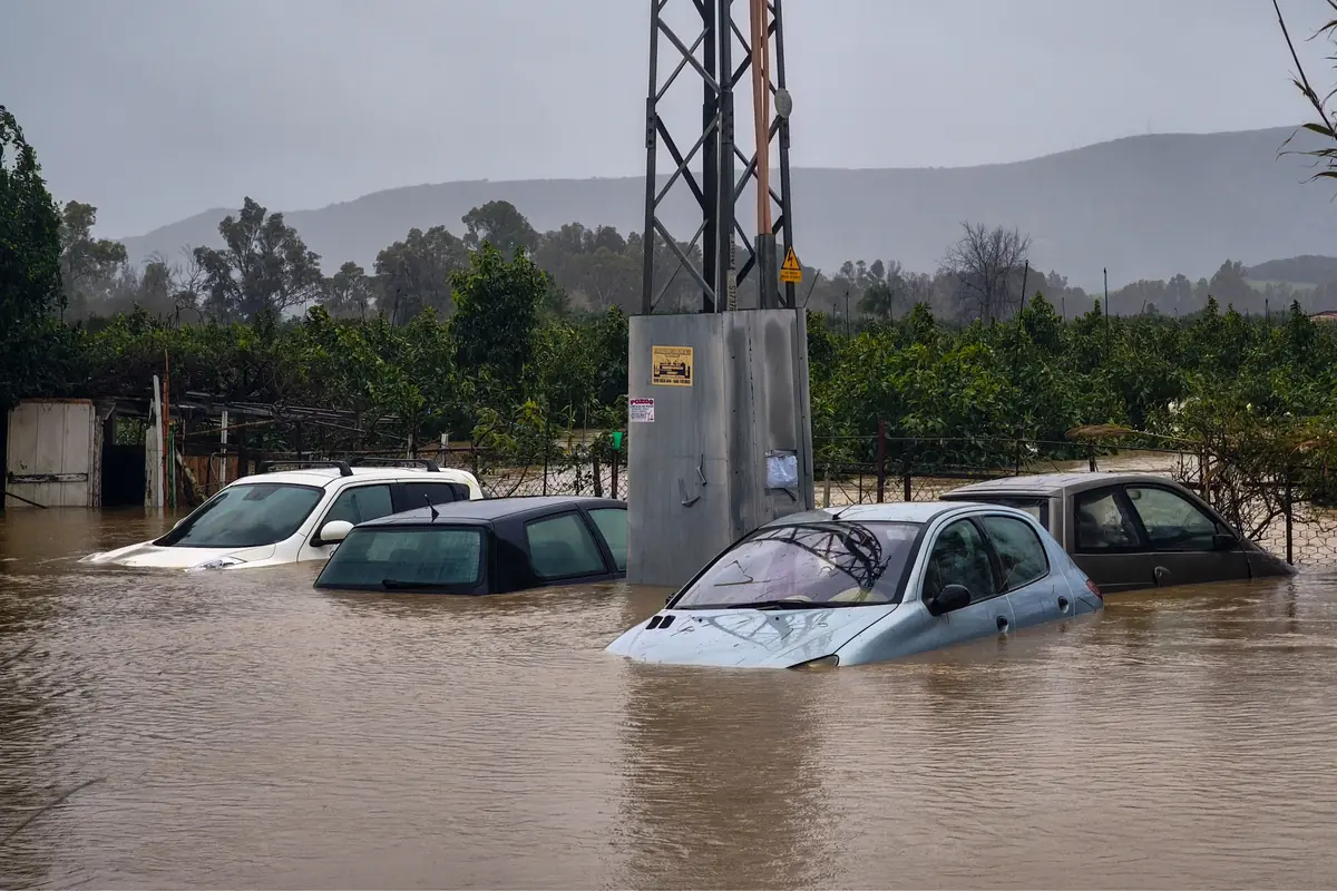 Coches casi sumergidos en una inundación junto a una torre eléctrica en una zona rural con árboles y montañas al fondo
