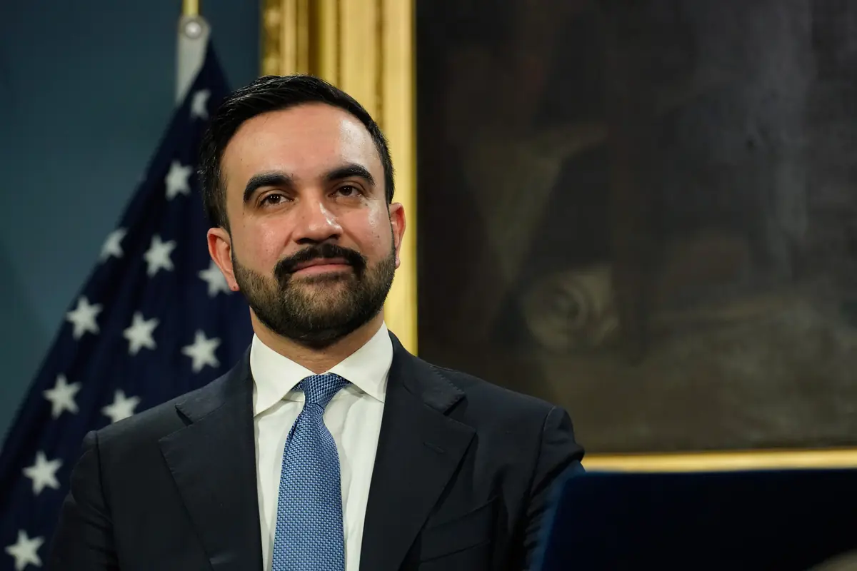 Hombre con barba y traje oscuro mirando al frente con una bandera de Estados Unidos desenfocada al fondo