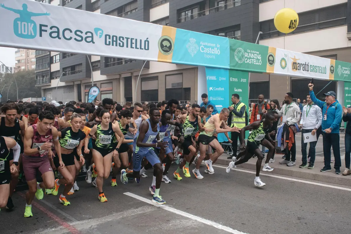 Salida multitudinaria de corredores en una carrera 10K urbana en Castellón bajo el arco de salida con público y organización observando desde los laterales Salida multitudinaria de corredores en una carrera 10K urbana en Castellón bajo el arco de salida con público y organización observando desde los laterales