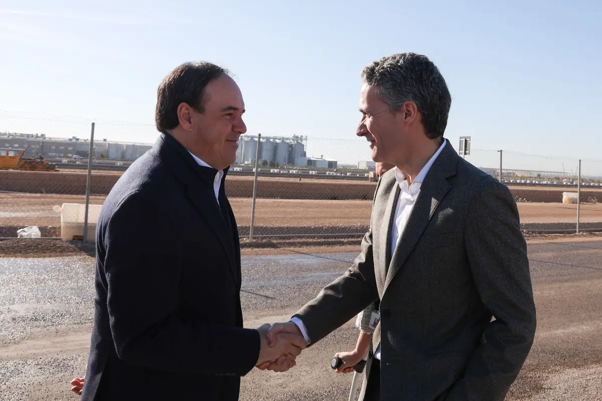Dos hombres de traje se dan la mano al aire libre junto a una obra cercada por vallas metálicas