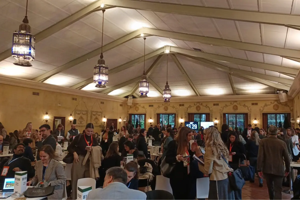 Salón amplio con techo a dos aguas lleno de personas conversando y trabajando en mesas durante un evento profesional o conferencia