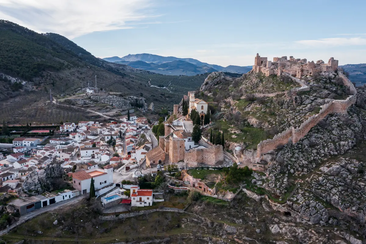 Vista aérea de un pueblo blanco andaluz junto a una colina rocosa coronada por un castillo amurallado medieval rodeado de montañas y olivares