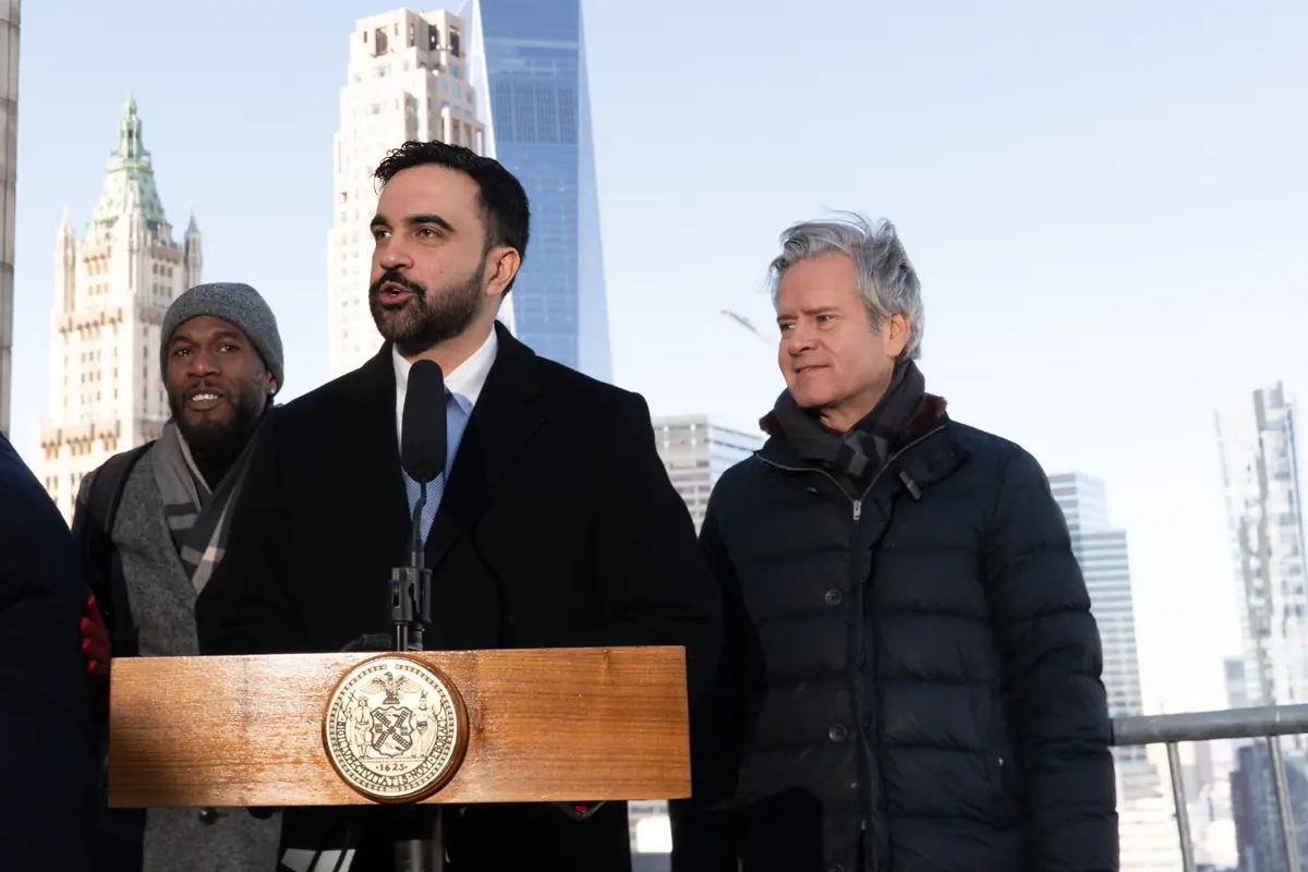 Hombre con barba hablando en un podio oficial al aire libre acompañado por otras dos personas con rascacielos de fondo Hombre con barba hablando en un podio oficial al aire libre acompañado por otras dos personas con rascacielos de fondo