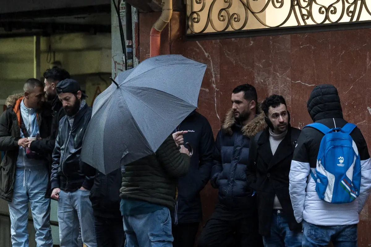 Grupo de hombres con ropa de abrigo esperando en la calle en un día lluvioso uno de ellos sostiene un paraguas gris