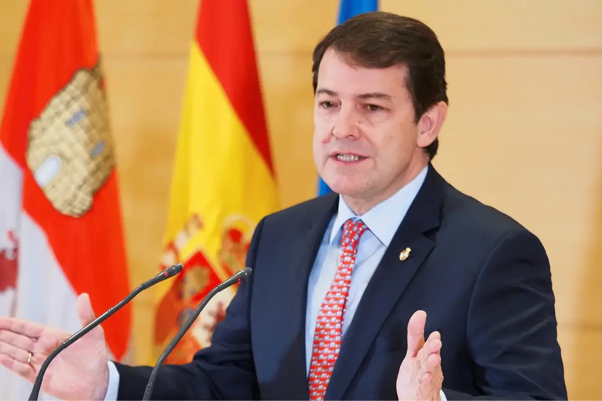 Hombre con traje y corbata hablando ante micrófonos con banderas de España y de una comunidad autónoma al fondo
