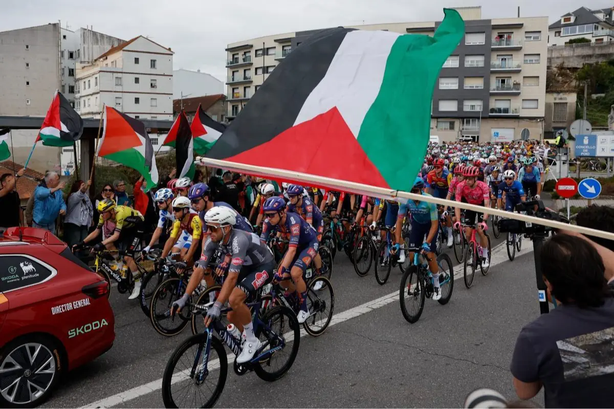 Pelotón de ciclistas compitiendo en carretera urbana mientras espectadores agitan grandes banderas palestinas en primera fila