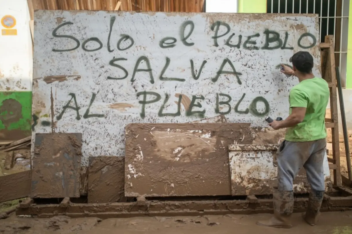 Hombre con botas embarradas escribe en una pared sucia el mensaje Solo el pueblo salva al pueblo en un entorno lleno de lodo