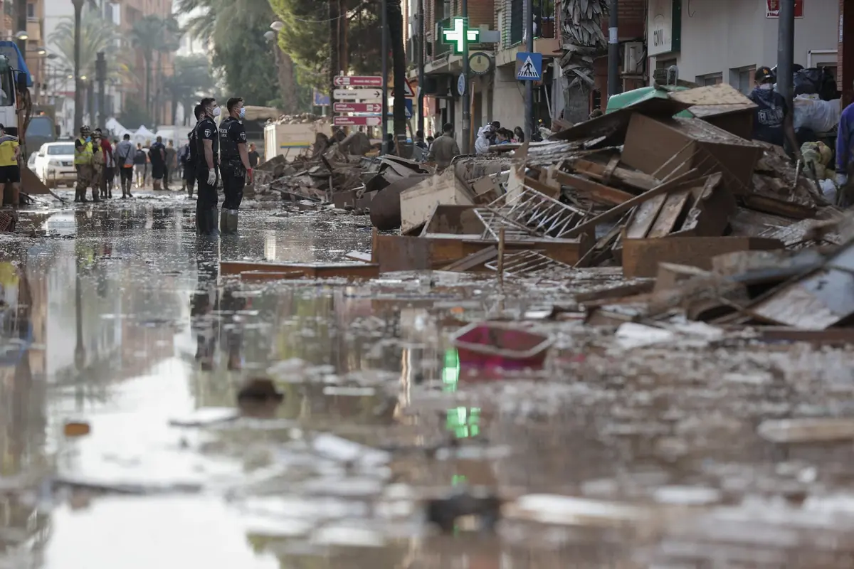 Calle urbana inundada con escombros y muebles amontonados mientras policías y equipos de emergencia recorren la zona tras una riada