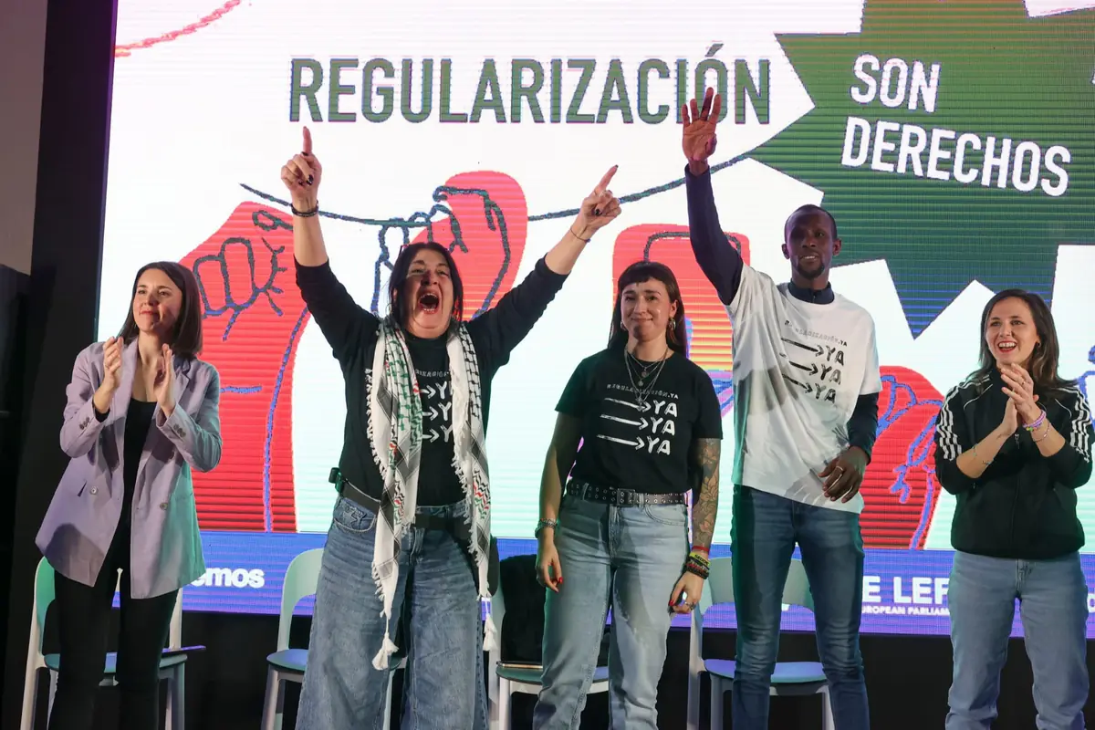 Grupo de personas de Podemos en un escenario celebrando y levantando los brazos frente a una pantalla con el mensaje Regularización son derechos