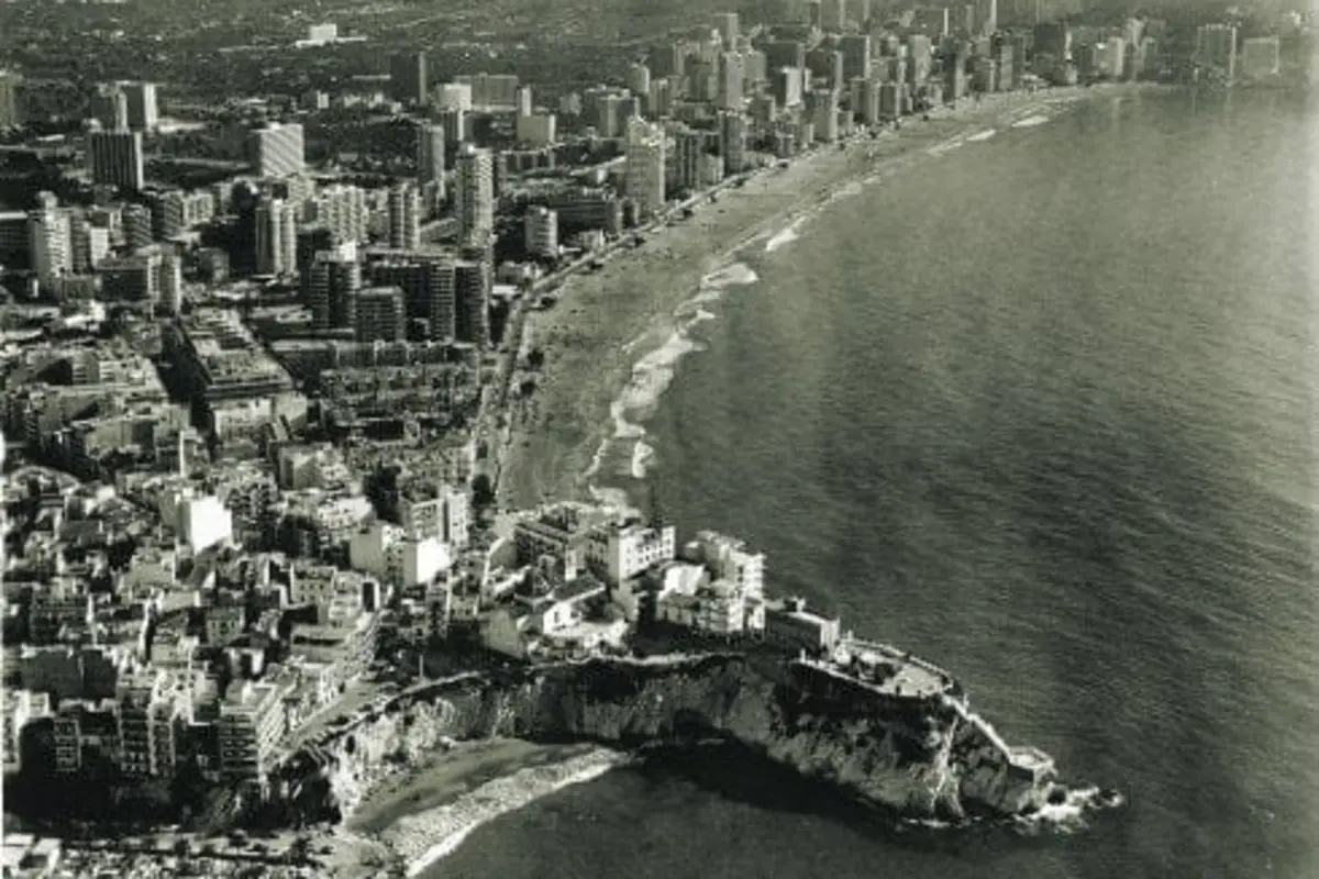 Vista aérea en blanco y negro de una ciudad costera con numerosos edificios altos junto a una larga playa curvada y un cabo rocoso que se adentra en el mar