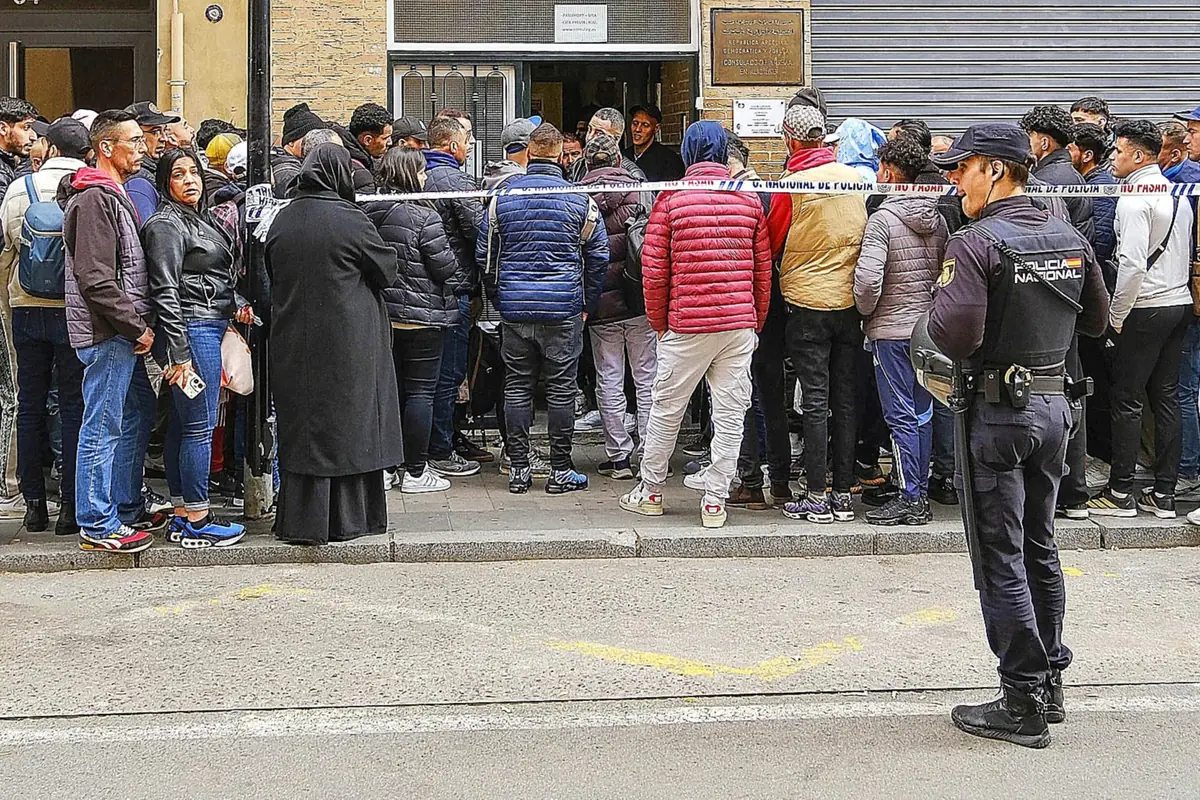 Multitud de personas haciendo fila en la acera frente a una oficina custodiada por una cinta policial y un agente de la Policía Nacional