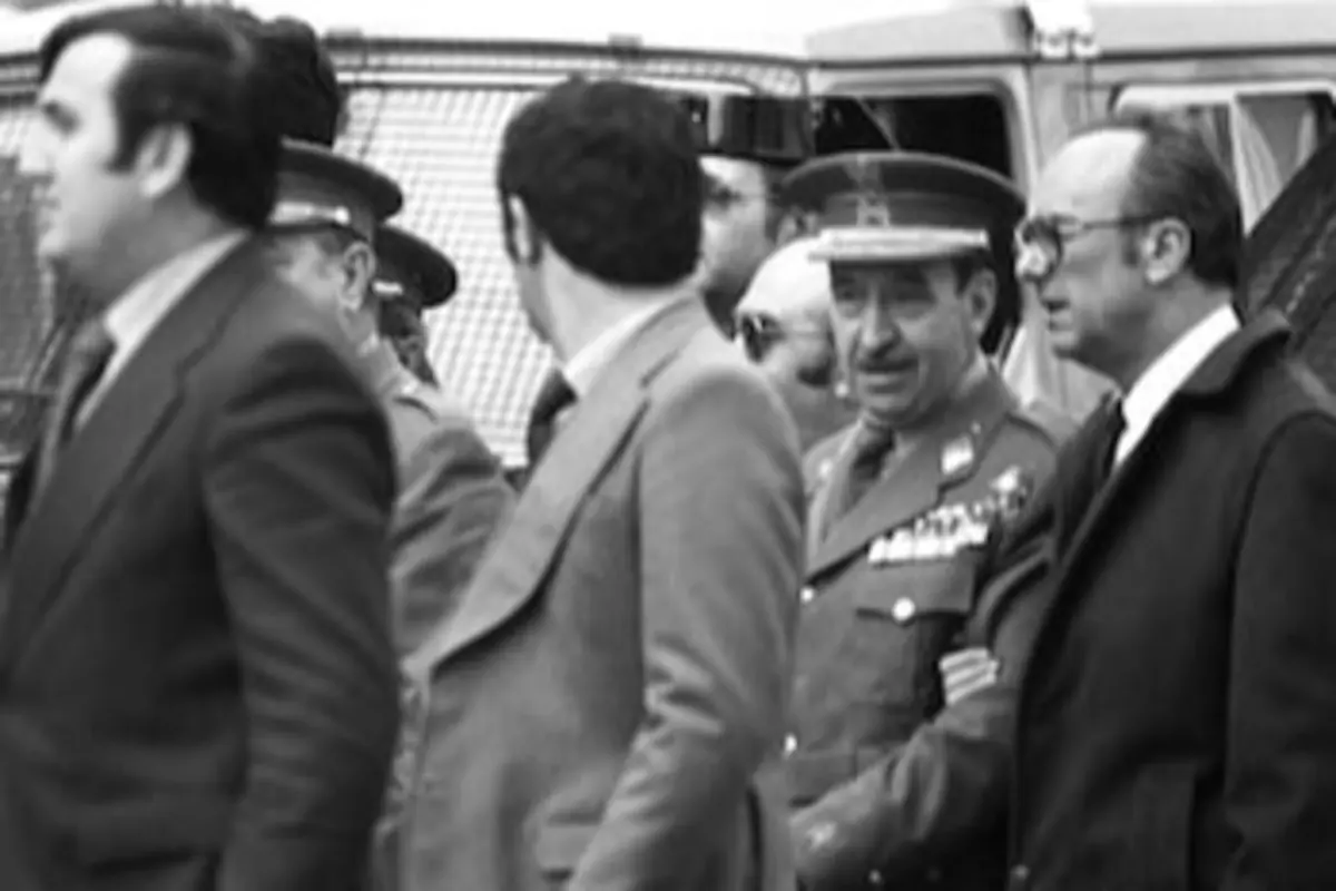 Fotografía en blanco y negro de varios hombres con traje y un militar con uniforme y medallas conversando en la calle frente a vehículos antiguos