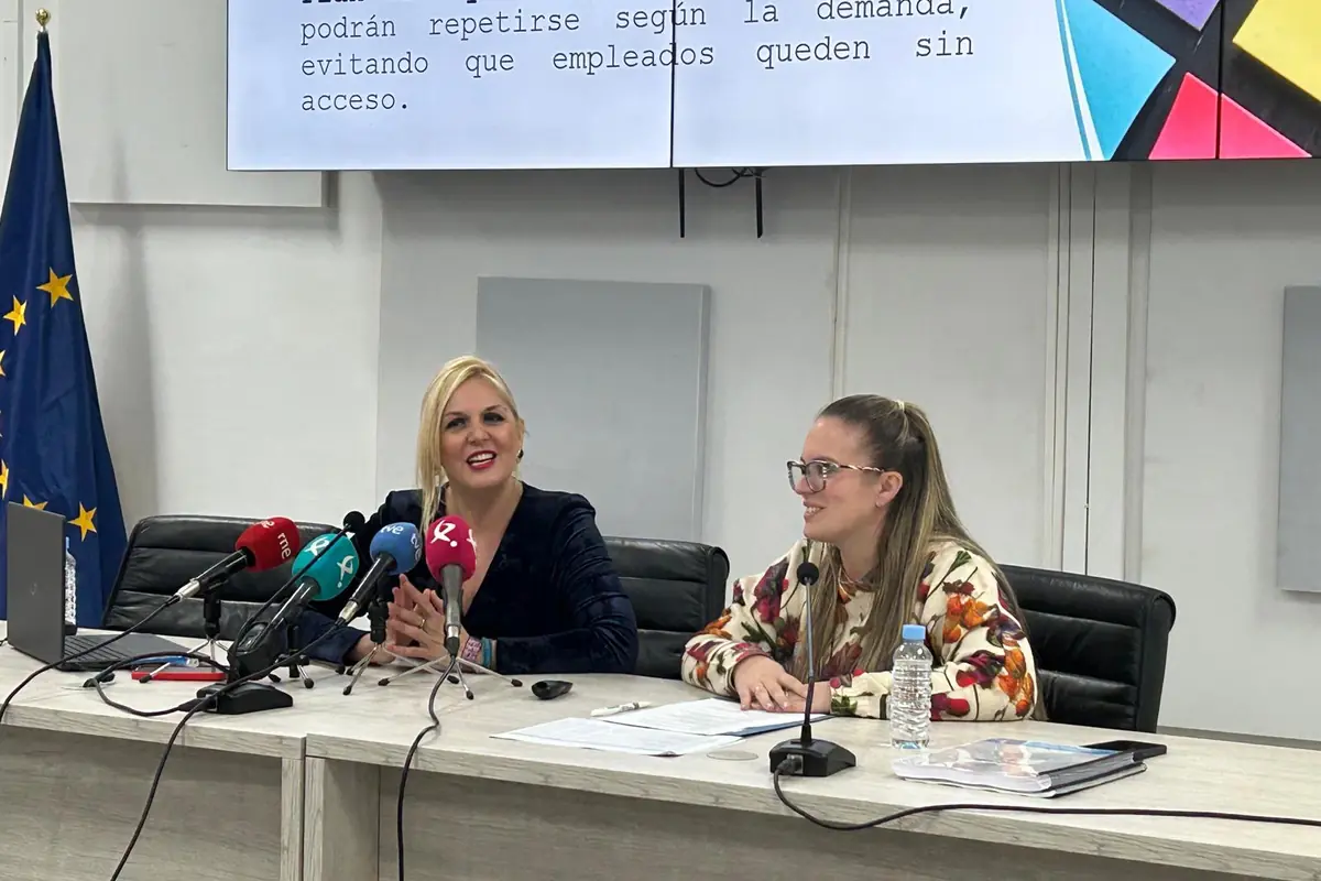 Dos mujeres sentadas en una mesa de conferencias con micrófonos y documentos frente a ellas mientras ofrecen una rueda de prensa en una sala institucional con bandera de la Unión Europea al fondo
