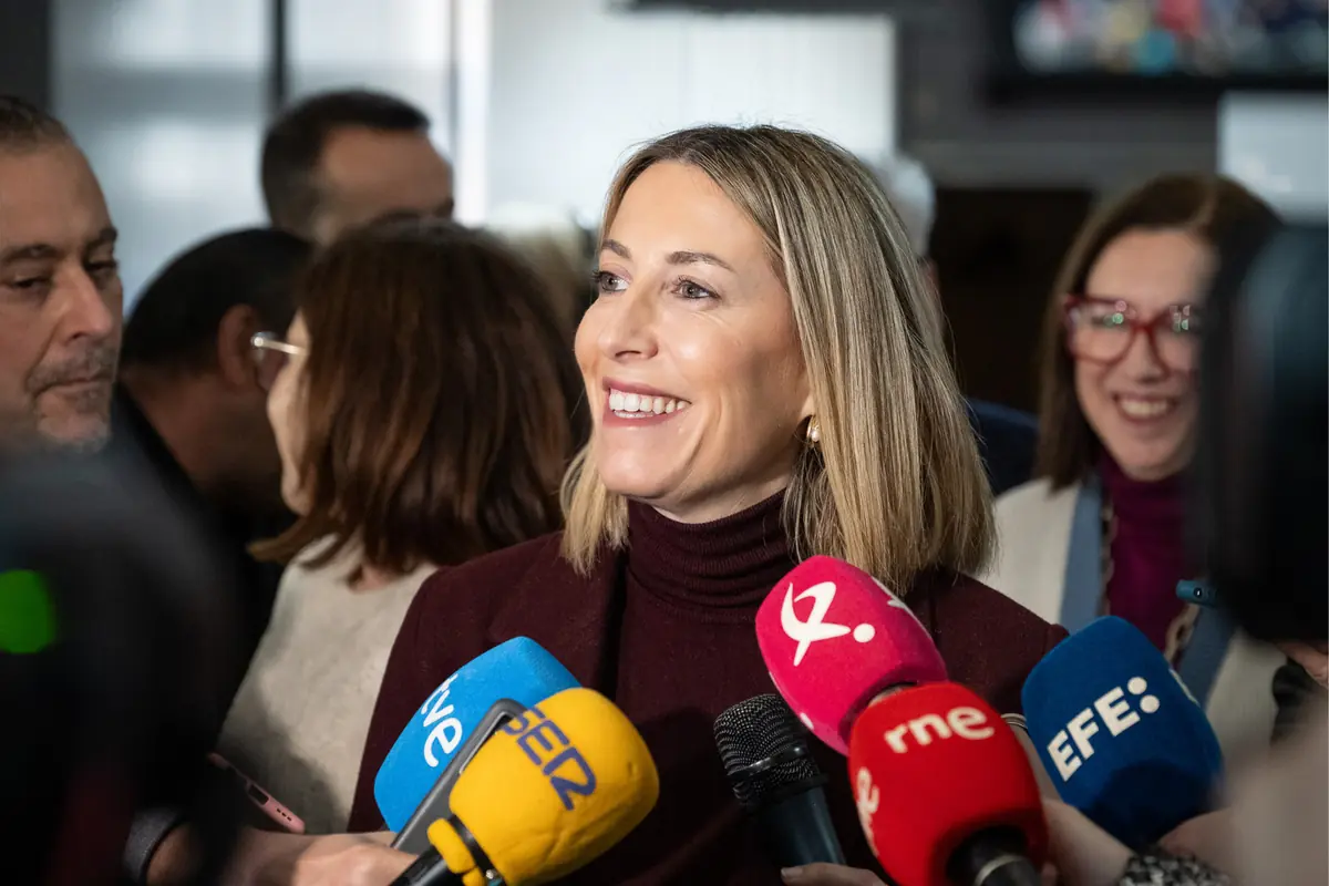 Mujer sonriendo mientras atiende a varios micrófonos de diferentes medios de comunicación en una rueda de prensa rodeada de periodistas Mujer sonriendo mientras atiende a varios micrófonos de diferentes medios de comunicación en una rueda de prensa rodeada de periodistas