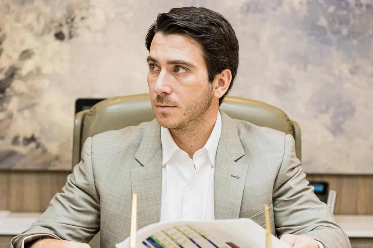 Hombre joven con traje claro sentado en una oficina mirando hacia un lado mientras sostiene documentos con gráficos de colores