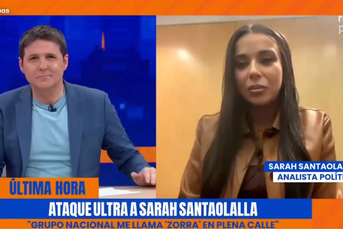Presentador de informativos en plató azul entrevista por videollamada a una analista política mientras un rótulo en pantalla anuncia un ataque ultra contra ella