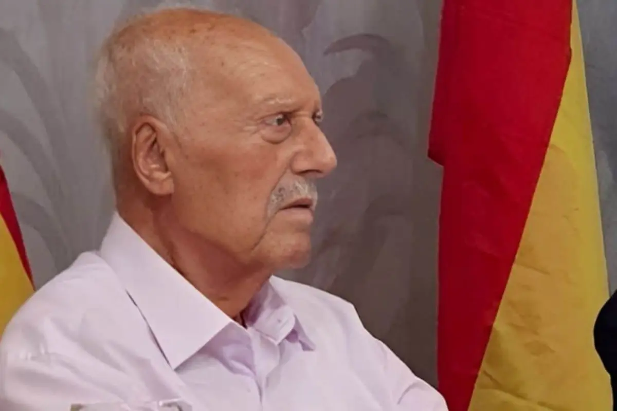 Hombre mayor de perfil con camisa clara sentado frente a una bandera roja y amarilla