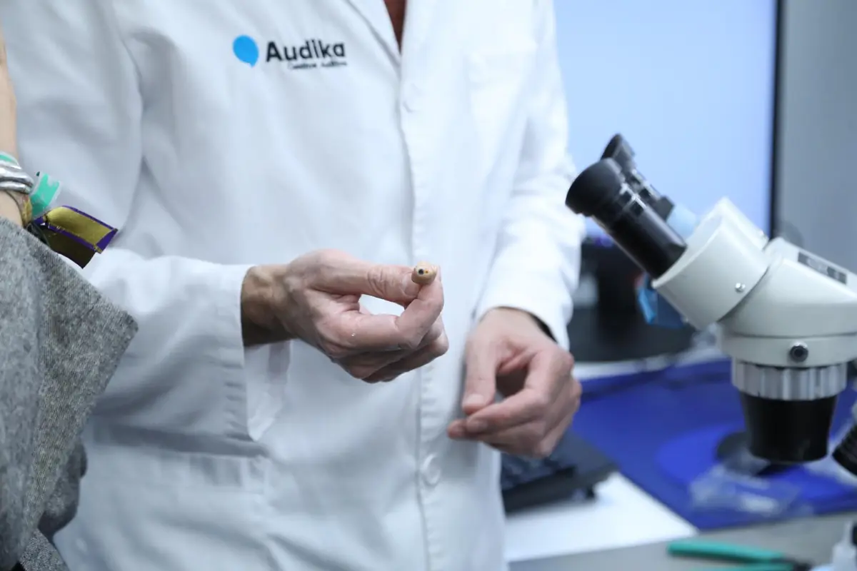 Profesional de la salud auditiva con bata blanca mostrando un pequeño audífono junto a un microscopio en un laboratorio