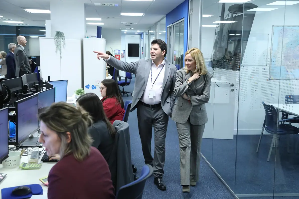 Dos personas con traje recorren una oficina moderna mientras varias empleadas trabajan en sus ordenadores