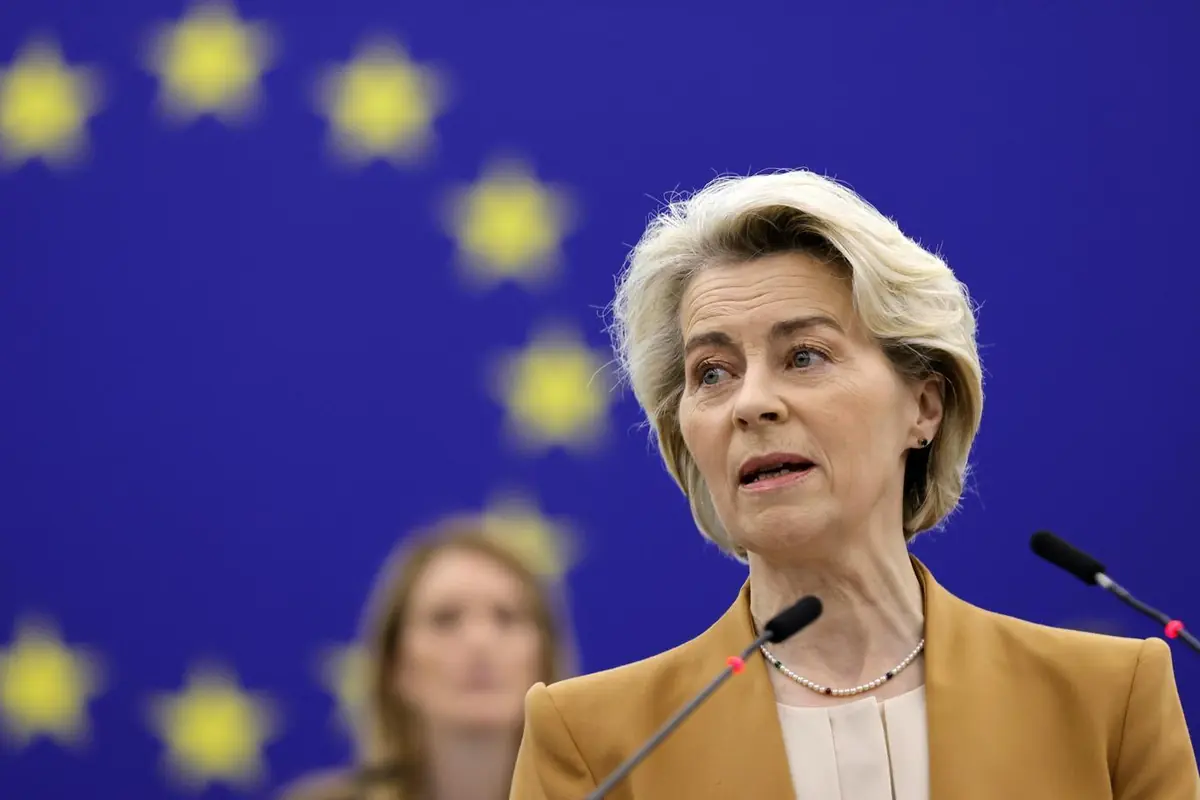Mujer de mediana edad hablando ante micrófonos con una gran bandera de la Unión Europea desenfocada al fondo