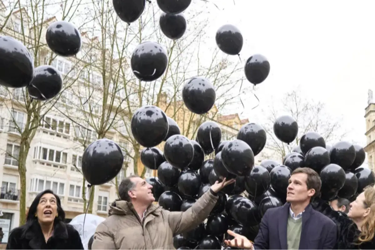 Personas en una plaza urbana sueltan numerosos globos negros que se elevan frente a edificios y árboles sin hojas