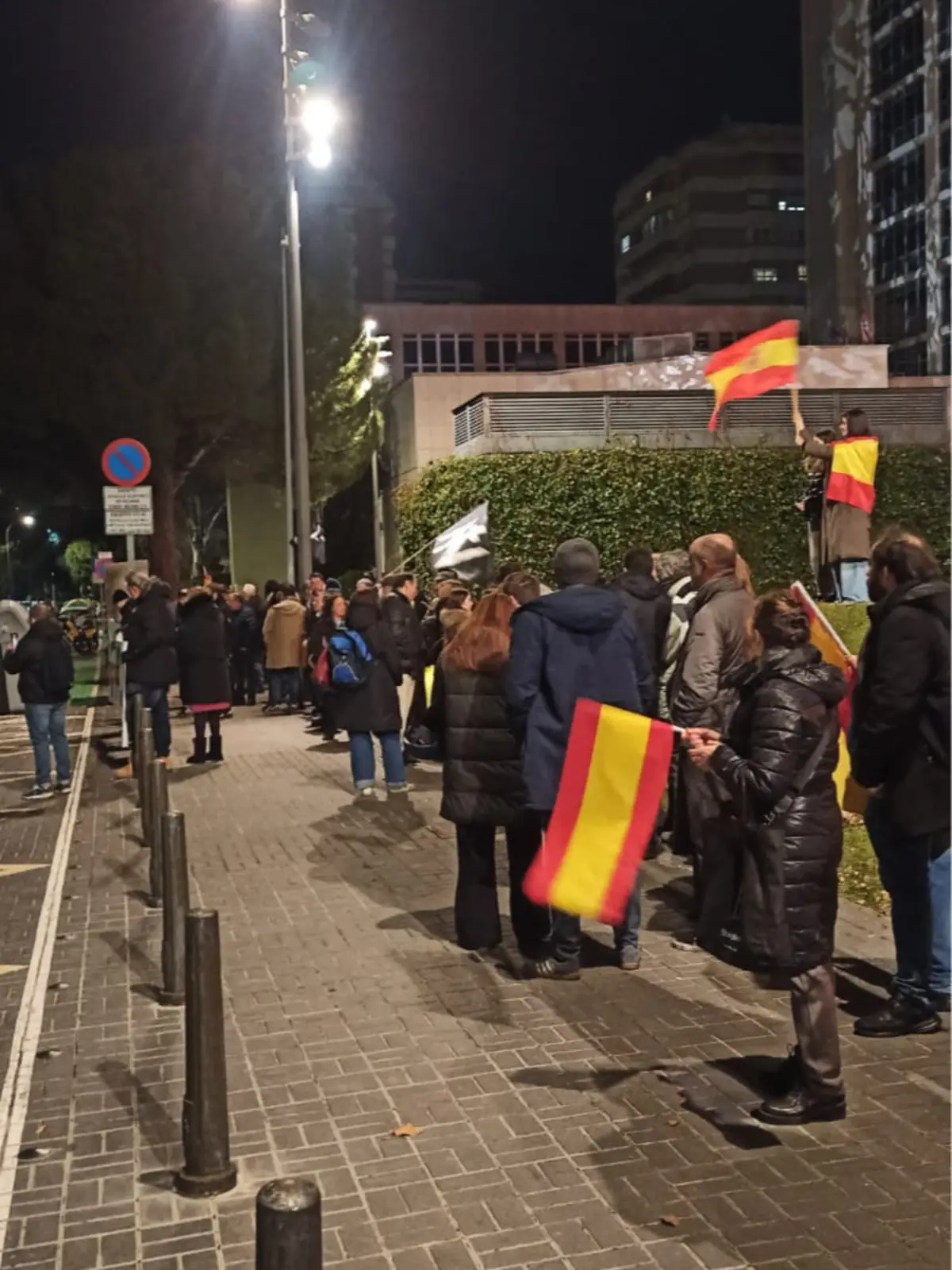 Grupo de personas manifestándose de noche en una calle urbana mientras varias de ellas sostienen banderas de España Grupo de personas manifestándose de noche en una calle urbana mientras varias de ellas sostienen banderas de España