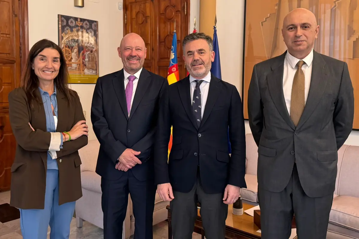 Cuatro personas posan de pie en una sala de reuniones elegante con sofás al fondo y banderas oficiales detrás de ellas