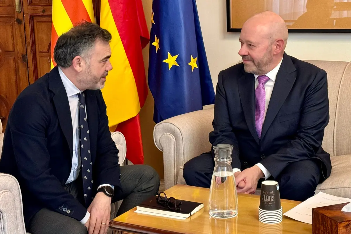 Dos hombres con traje conversan sentados en un despacho oficial con banderas de España Cataluña y la Unión Europea al fondo y una mesa con documentos agua y vasos de café delante