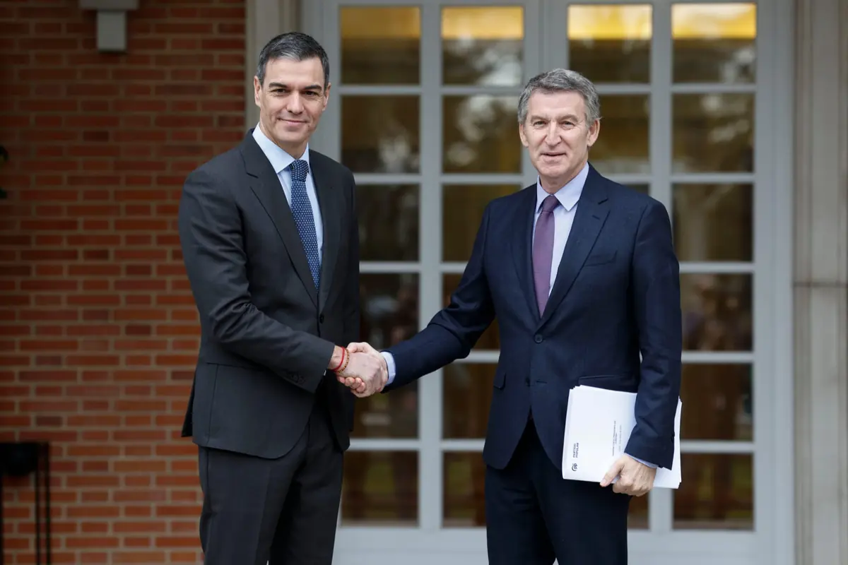 Dos hombres con traje oscuro se dan la mano frente a la entrada de un edificio con pared de ladrillo y puerta acristalada