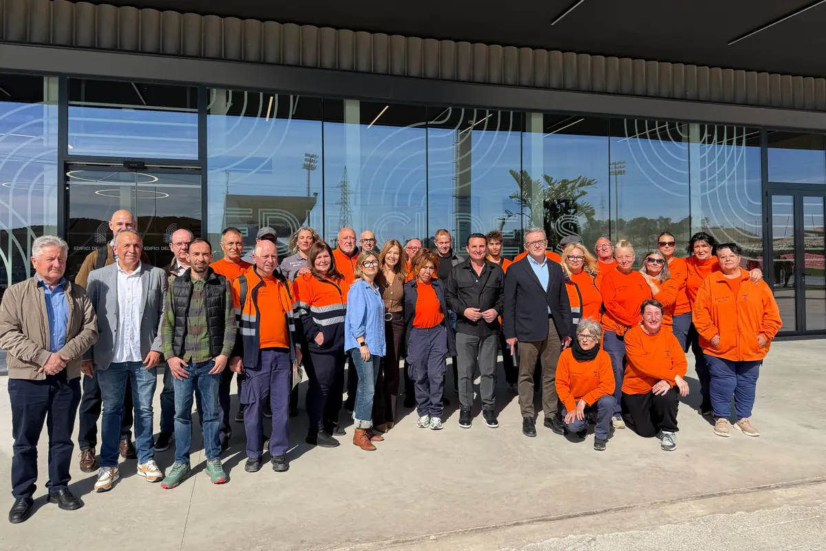 Grupo numeroso de trabajadores y trabajadoras con uniformes naranjas y ropa casual posando sonrientes frente a la entrada acristalada de un edificio moderno