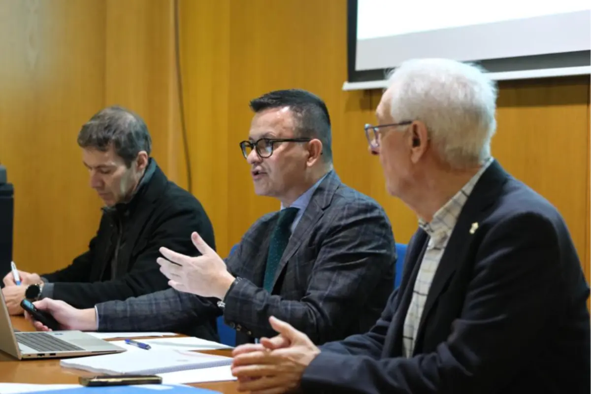 Tres hombres sentados en una mesa de reuniones mientras uno de ellos habla y gesticula durante una presentación con documentos y un portátil delante