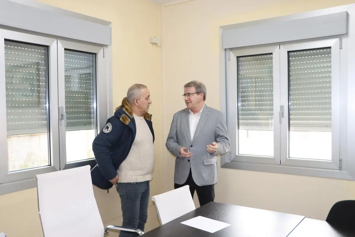 Dos hombres conversan de pie junto a una mesa en una oficina luminosa con grandes ventanas y persianas bajadas