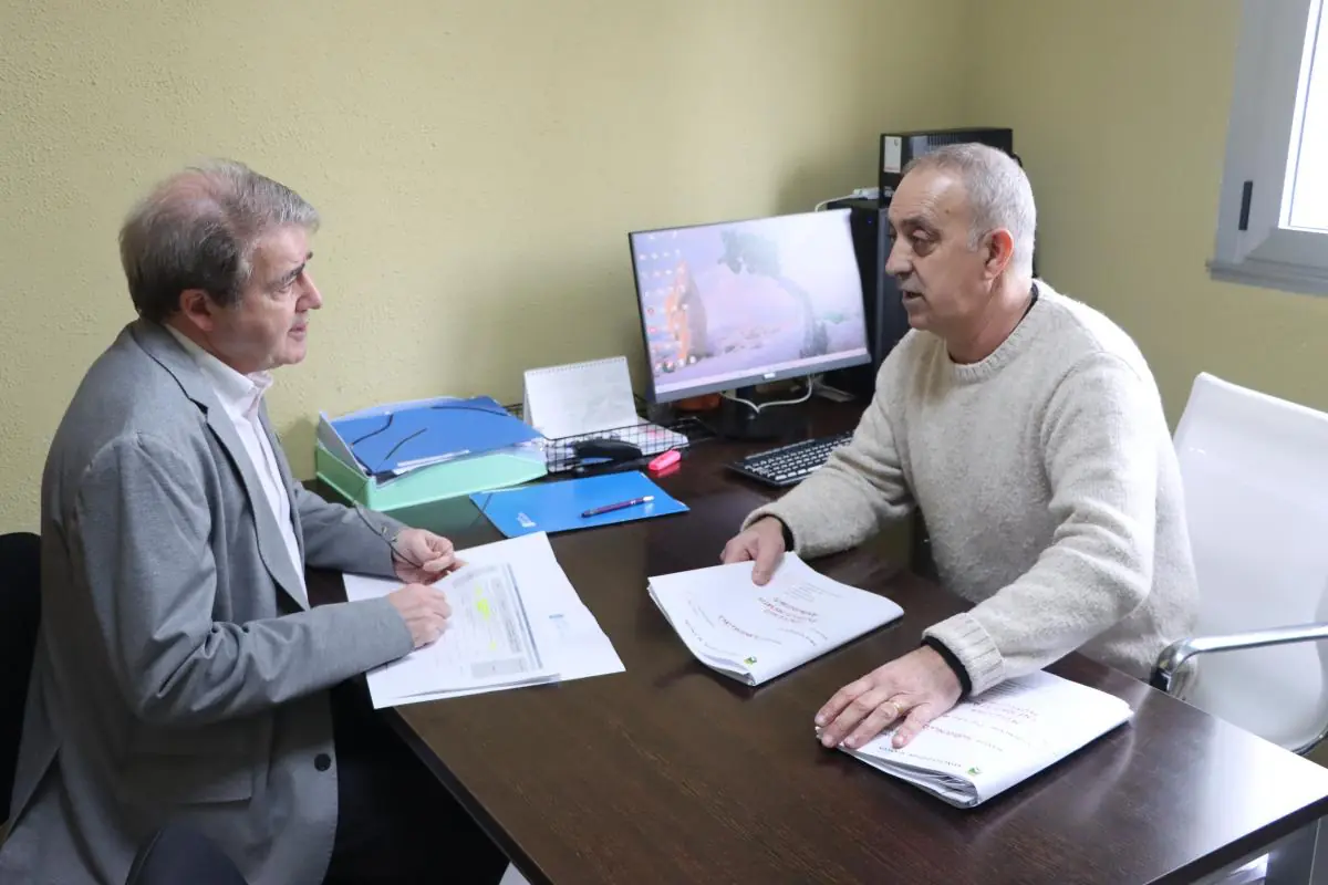 Dos hombres maduros conversan en una oficina sentados frente a frente en un escritorio con documentos y un ordenador encendido