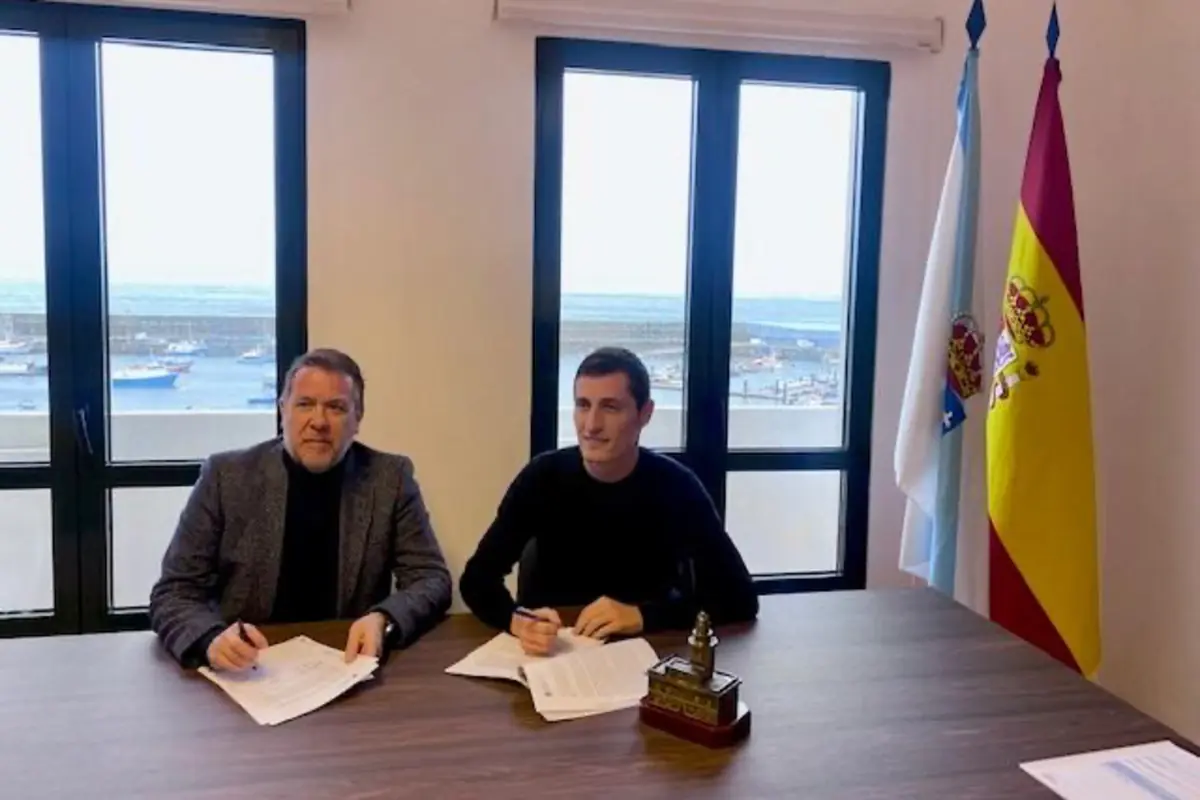 Dos hombres sentados en una mesa firmando documentos en una oficina con vista al puerto y banderas oficiales de España y de una comunidad autónoma al fondo